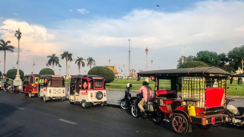 Open-sided tuk-tuks and motorized golf carts wait for fares outside Royal Palace Park. 