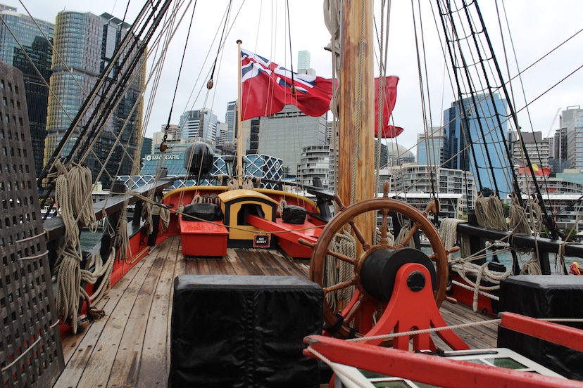 On the deck of the HMB Endeavour replica with its rope ladders and rigging, moored at the Australian National Maritime Museum in Sydney. At the ship’s stern is the Australian Red Ensign maritime flag.